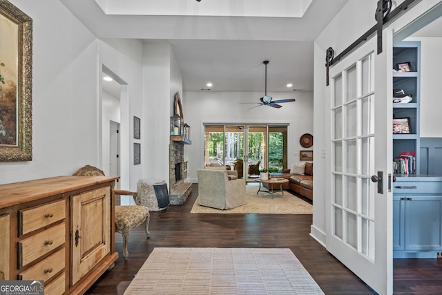 living room with built in features, ceiling fan, dark hardwood / wood-style floors, a stone fireplace, and a barn door