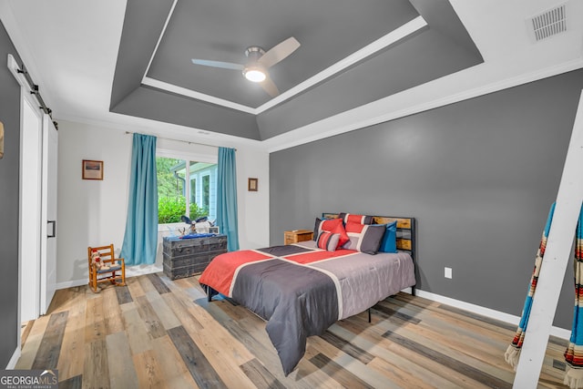 bedroom with hardwood / wood-style floors, ornamental molding, ceiling fan, a raised ceiling, and a barn door