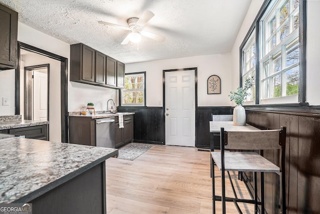 kitchen with stainless steel dishwasher, a wainscoted wall, ceiling fan, light wood-type flooring, and a textured ceiling