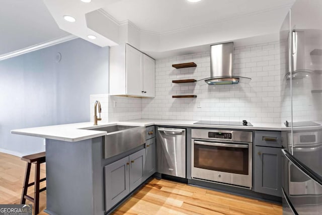 kitchen with wall chimney range hood, gray cabinetry, stainless steel appliances, a kitchen bar, and kitchen peninsula