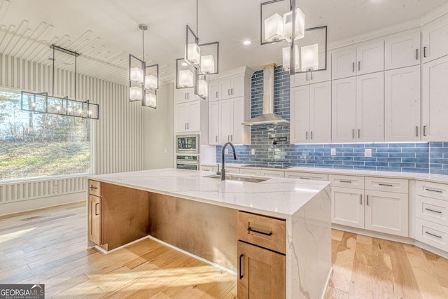 kitchen with white cabinetry, oven, a center island with sink, light stone countertops, and wall chimney range hood