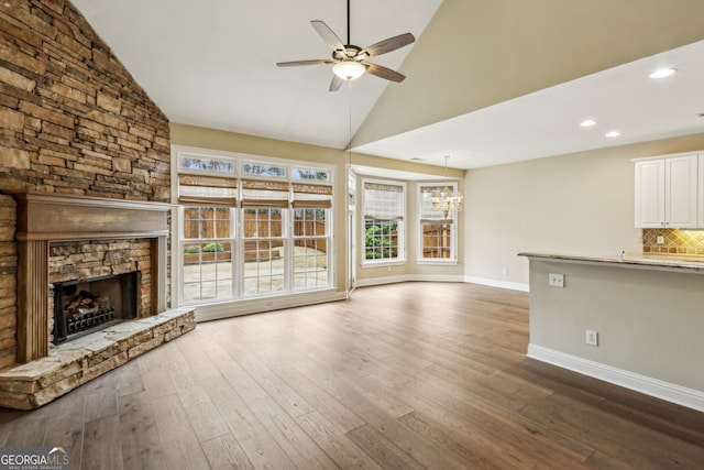 unfurnished living room with a stone fireplace, ceiling fan with notable chandelier, high vaulted ceiling, and dark hardwood / wood-style floors