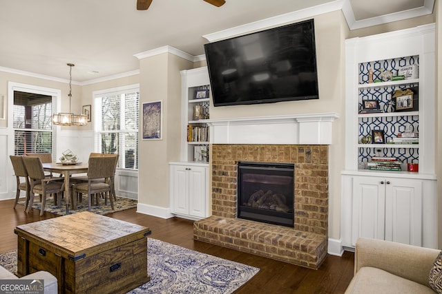 living room with dark wood-type flooring, ceiling fan with notable chandelier, ornamental molding, a brick fireplace, and built in features