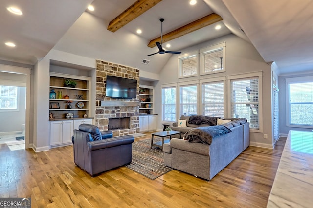 living room featuring built in shelves, a stone fireplace, ceiling fan, beam ceiling, and light hardwood / wood-style floors