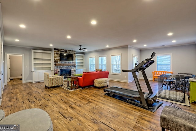 workout area featuring built in features, ceiling fan, wood-type flooring, ornamental molding, and a stone fireplace