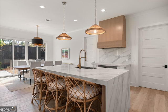 kitchen featuring an island with sink, sink, decorative backsplash, hanging light fixtures, and light stone counters