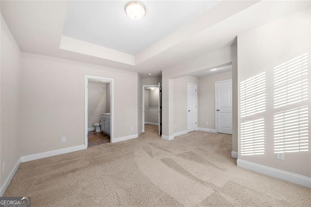 unfurnished bedroom featuring a tray ceiling, a walk in closet, light colored carpet, and ensuite bathroom