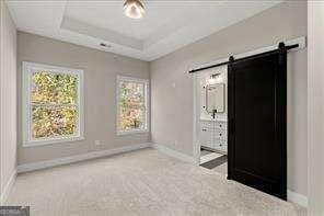 unfurnished bedroom with ensuite bathroom, a tray ceiling, a barn door, and carpet