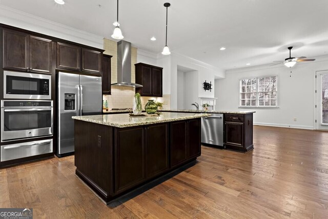 kitchen with appliances with stainless steel finishes, a kitchen island, wall chimney exhaust hood, hanging light fixtures, and dark brown cabinets