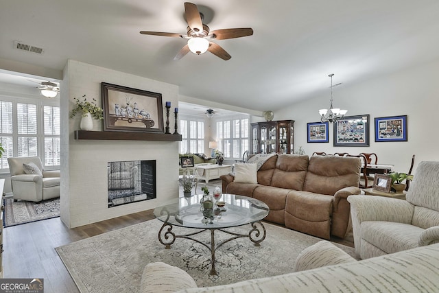 living area with lofted ceiling, ceiling fan with notable chandelier, wood finished floors, visible vents, and a brick fireplace