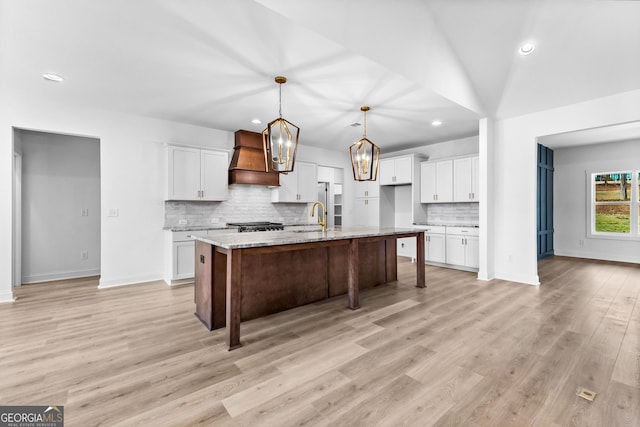 kitchen featuring premium range hood, decorative light fixtures, an island with sink, white cabinets, and light stone countertops