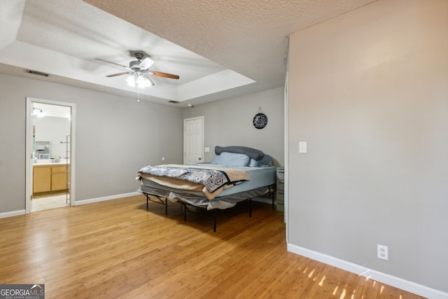 bedroom featuring light hardwood / wood-style flooring, ceiling fan, connected bathroom, a textured ceiling, and a raised ceiling