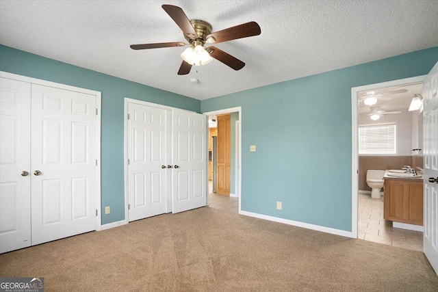 unfurnished bedroom with ceiling fan, ensuite bathroom, a textured ceiling, two closets, and light colored carpet