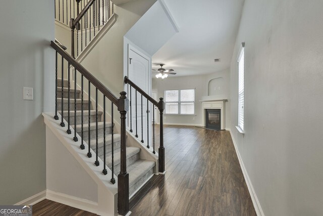 stairway featuring wood finished floors, visible vents, a ceiling fan, baseboards, and a glass covered fireplace