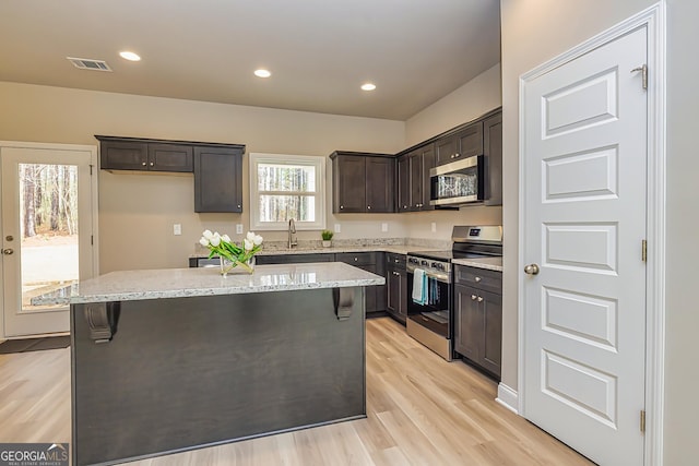 kitchen with dark brown cabinetry, light stone counters, a center island, stainless steel appliances, and light hardwood / wood-style floors