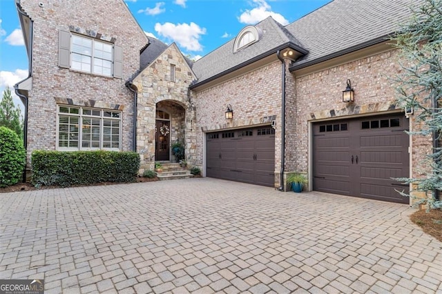 french country inspired facade featuring decorative driveway, brick siding, and an attached garage