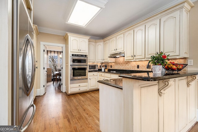 kitchen with dark countertops, a peninsula, stainless steel appliances, cream cabinetry, and under cabinet range hood