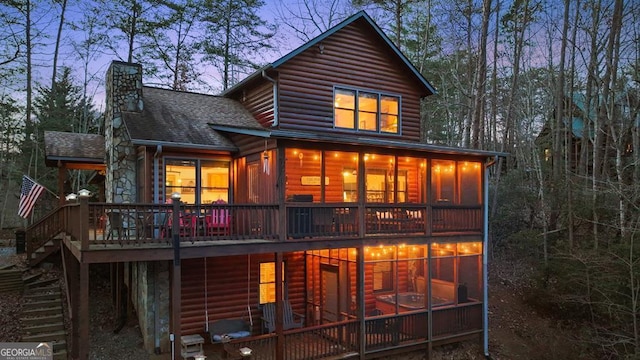 rear view of house with a shingled roof, faux log siding, a sunroom, a chimney, and stairs