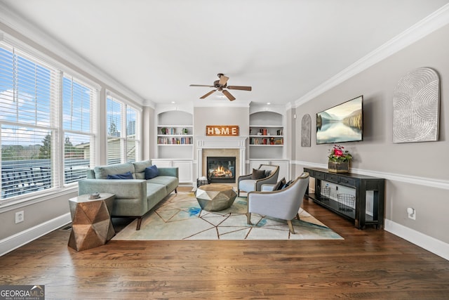 living room with built in shelves, wood finished floors, baseboards, a glass covered fireplace, and crown molding