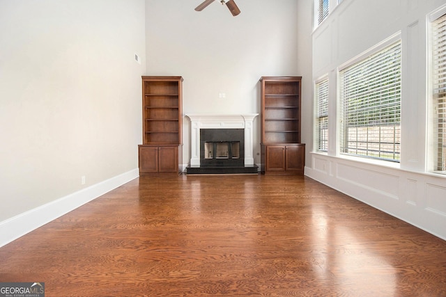 unfurnished living room featuring a towering ceiling, dark wood-style flooring, a fireplace with raised hearth, ceiling fan, and healthy amount of natural light