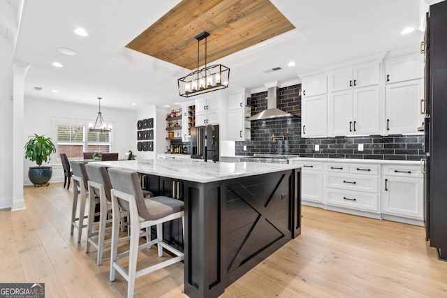 kitchen featuring a raised ceiling, white cabinetry, a kitchen island with sink, and wall chimney exhaust hood