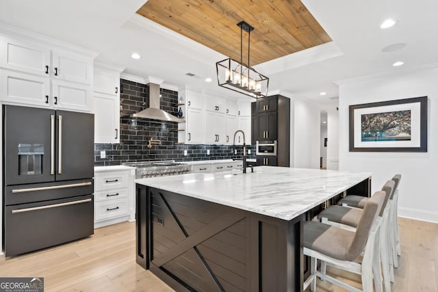 kitchen with appliances with stainless steel finishes, an island with sink, white cabinets, a tray ceiling, and wall chimney exhaust hood