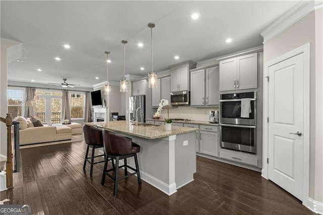 kitchen with decorative light fixtures, gray cabinetry, stainless steel appliances, light stone countertops, and a center island with sink