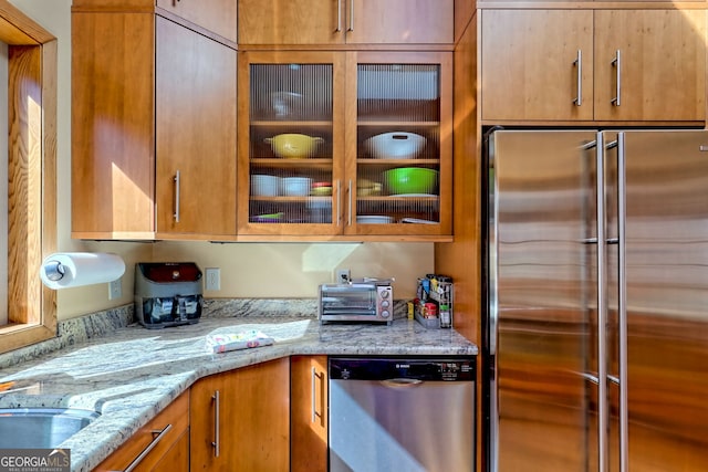 kitchen featuring a toaster, brown cabinetry, glass insert cabinets, light stone counters, and stainless steel appliances