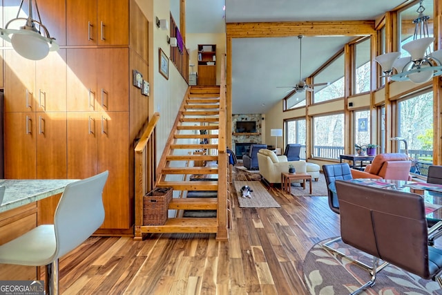 living area featuring ceiling fan, high vaulted ceiling, a stone fireplace, beam ceiling, and light wood finished floors