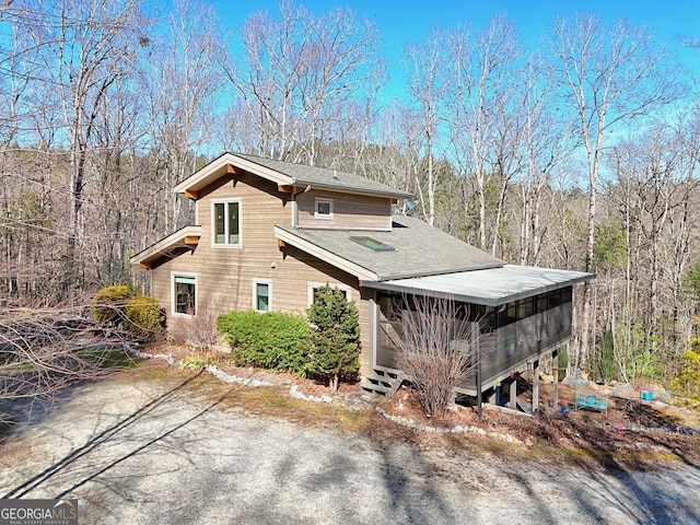 view of property exterior featuring a sunroom, a shingled roof, and a view of trees