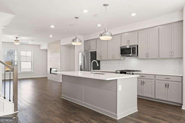 kitchen with gray cabinetry, hanging light fixtures, backsplash, stainless steel appliances, and an island with sink