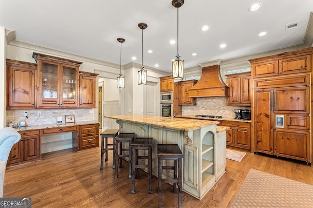 kitchen with custom exhaust hood, built in appliances, hanging light fixtures, a kitchen island, and light stone countertops