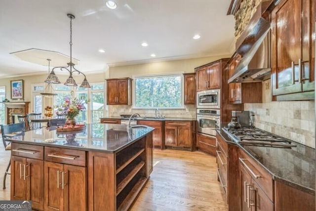 kitchen featuring a kitchen island with sink, stainless steel appliances, decorative light fixtures, range hood, and crown molding