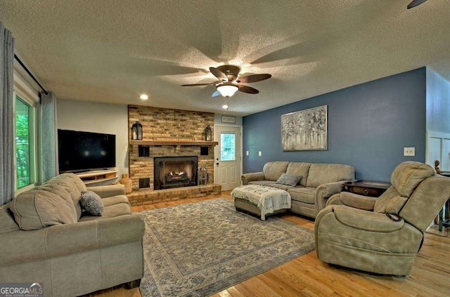 living room featuring a brick fireplace, a textured ceiling, light hardwood / wood-style flooring, and ceiling fan