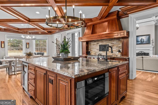 kitchen with sink, a center island with sink, coffered ceiling, a notable chandelier, and stone countertops