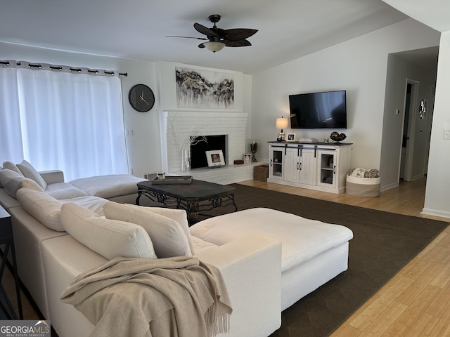 living room featuring baseboards, vaulted ceiling, a brick fireplace, wood finished floors, and a ceiling fan