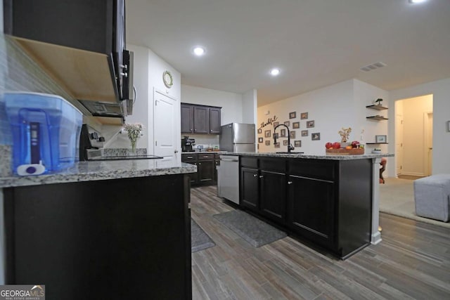 kitchen featuring stainless steel appliances, light stone countertops, an island with sink, and wood-type flooring