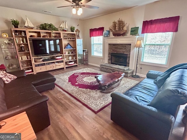 living area featuring a stone fireplace, wood finished floors, baseboards, and ceiling fan