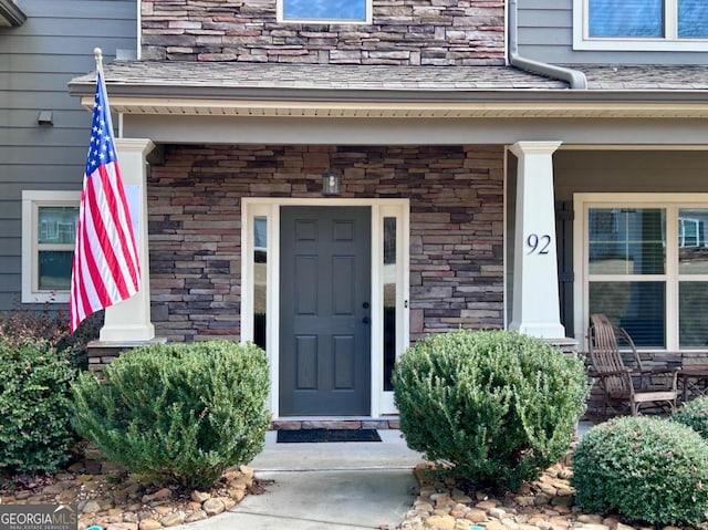 entrance to property with covered porch, stone siding, and roof with shingles