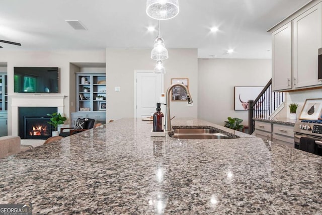 kitchen with stainless steel gas range oven, dark stone counters, a lit fireplace, pendant lighting, and a sink