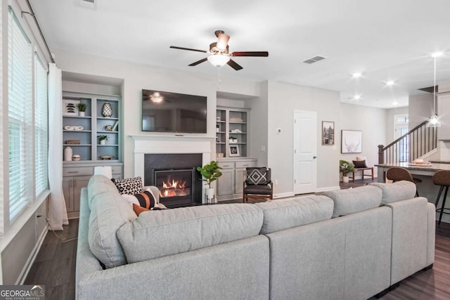 living area featuring built in shelves, dark wood-type flooring, visible vents, stairs, and a glass covered fireplace