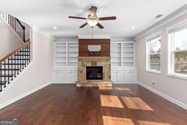 unfurnished living room featuring built in shelves, a stone fireplace, ceiling fan, crown molding, and dark hardwood / wood-style floors