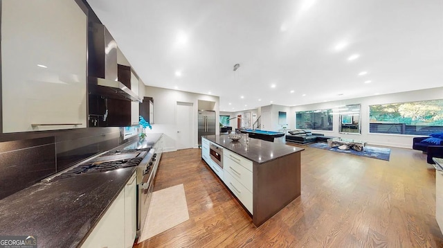 kitchen featuring wall chimney exhaust hood, wood finished floors, open floor plan, and built in appliances