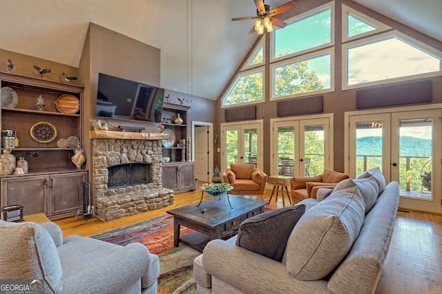 living room featuring a stone fireplace, high vaulted ceiling, ceiling fan, light wood-type flooring, and french doors