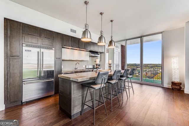 kitchen featuring hanging light fixtures, a breakfast bar area, built in fridge, expansive windows, and dark hardwood / wood-style flooring