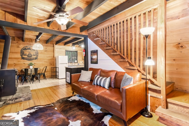 living room featuring a wood stove, beam ceiling, wood walls, light hardwood / wood-style floors, and ceiling fan