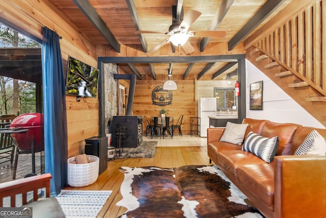 living room with wood-type flooring, a wood stove, ceiling fan, wooden walls, and beam ceiling