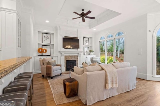 living room with built in shelves, light hardwood / wood-style flooring, ornamental molding, a raised ceiling, and ceiling fan