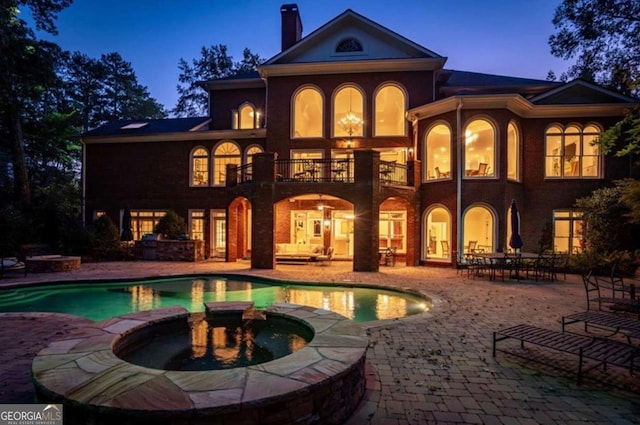 back house at dusk featuring a swimming pool with hot tub, a patio area, and a balcony