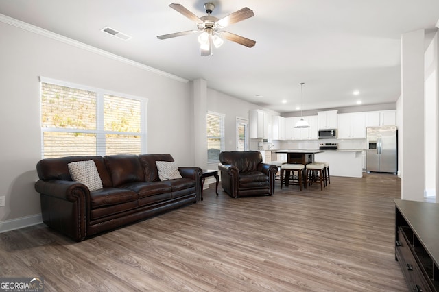 living area with ceiling fan, wood finished floors, visible vents, baseboards, and crown molding